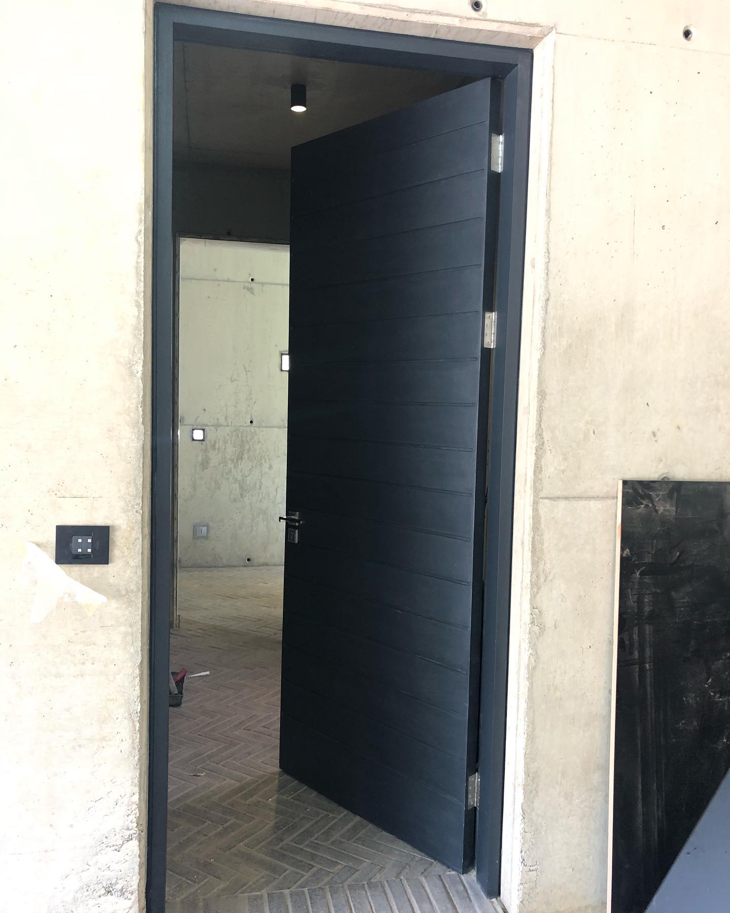 Black horizontal-slat pivot door standing open against a raw concrete surround